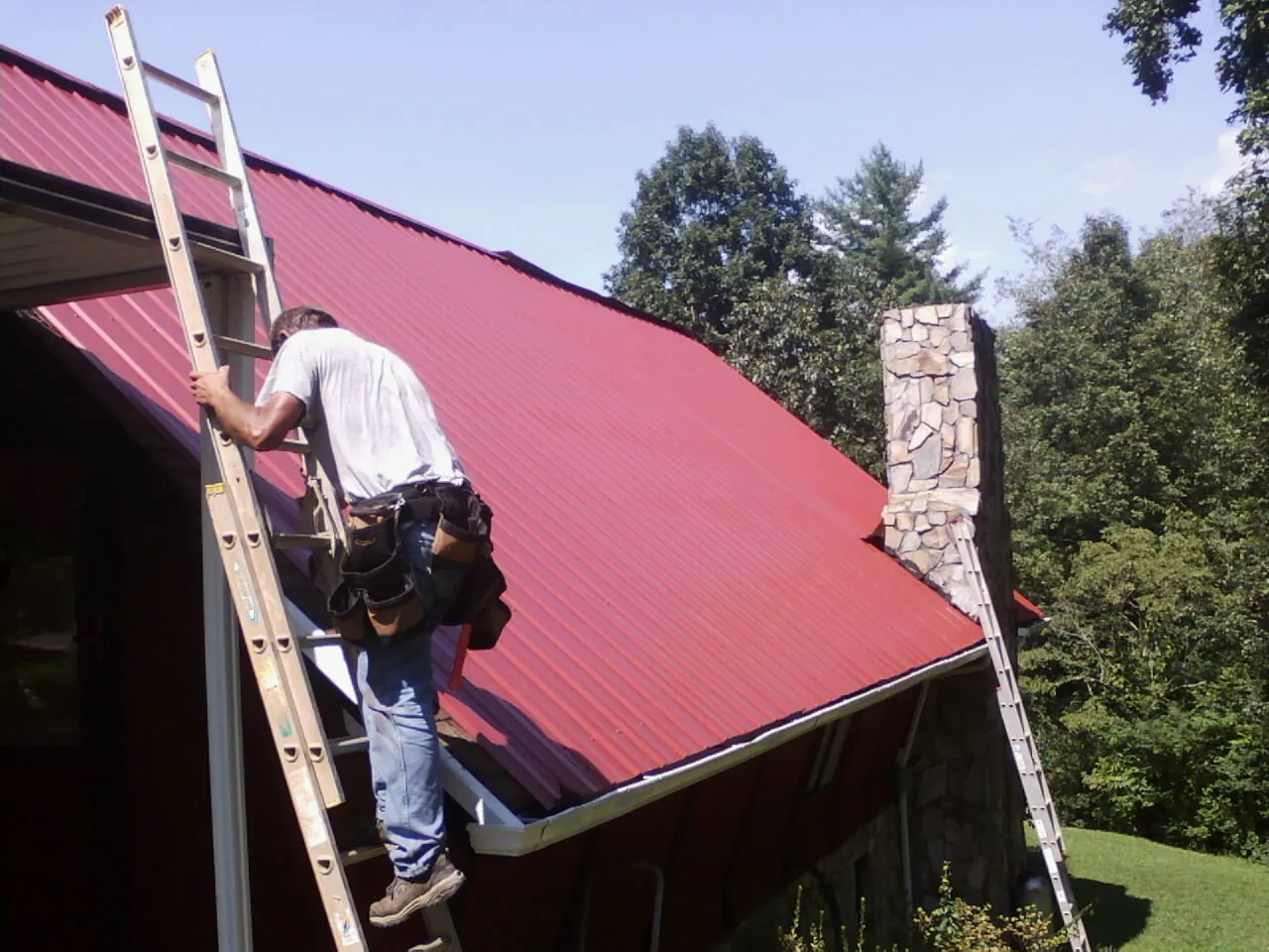Roofer on a ladder beside a red metal roof and stone chimney — Soffit & Fascia Repair by Roofing Brothers in Mc Grady